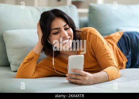 Portrait Of Smiling Arab Woman Wearing Earphones Relaxing With Smartphone At Home Stock Photo