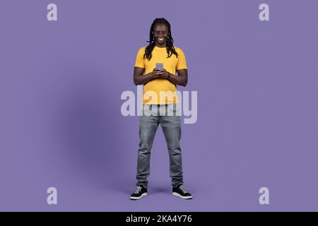 Happy stylish black guy using smartphone on purple, full length Stock Photo