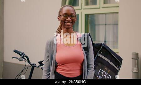 Young smiling courier waiting to customer outdoors, standing next to bike to deliver restaurant meal order in thermal bag. Carrying backpack and holding fast food, express services. Stock Photo