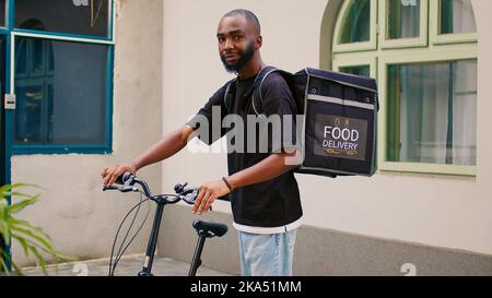 Happy african american courier waiting for client outside, riding bicycle with thermal backpack to deliver takeaway meal. Deliveryman standing near office building entrance, looking at camera. Stock Photo