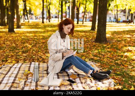 Concentrated smart young european red-haired lady student in raincoat sit with phone makes notes in diary Stock Photo