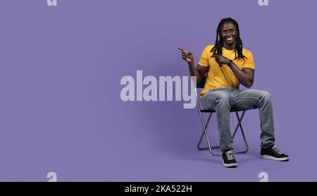 Relaxed black man sitting on chair, pointing at copy space Stock Photo