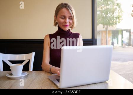 Beautiful mature blonde woman happily telecommuting from coffee shop, using laptop. Stock Photo