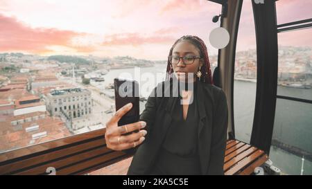 Portrait of a cute young black female tourist in the car of a ropeway taking a selfie on her cellphone; a youthful African woman is having a video cal Stock Photo