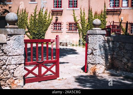 An outdoor shot of an entrance to the local area with a selective focus on the closed leaf of the red wooden gate surrounded by stone walls on a warm Stock Photo