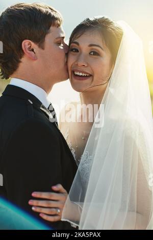 I can get use to this. Portrait of a cheerful young groom giving his bride a kiss on the cheek while they stand holding each other outside. Stock Photo
