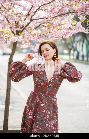 A spectacular woman in a bright dress stands against the background of sakura. A dark-haired woman in a beautiful outfit smiles on the street while wa Stock Photo