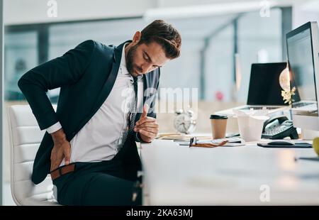 Working until your back hurts. a uncomfortable young businessman holding his back in discomfort due to pain while being sated in the office. Stock Photo