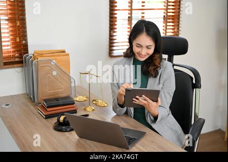 Professional millennial Asian female lawyer or business legal consultant using digital tablet while sitting at her desk in modern office. Stock Photo