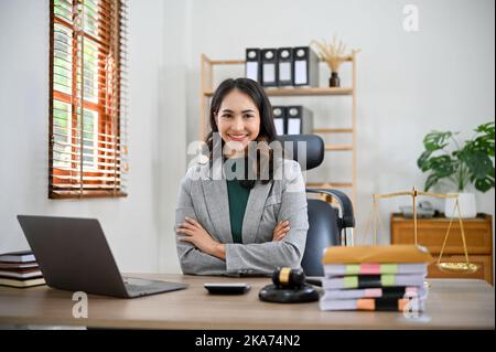 Professional and successful millennial Asian female lawyer or attorney in formal suit, arms crossed, sitting at her desk, smiling and looking at the c Stock Photo
