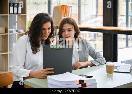 Pretty and charming young caucasian businesswoman looking at the details on clipboard, discussing work with her coworker in the office. Stock Photo