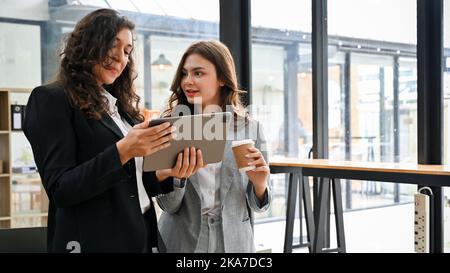 Professional millennial Caucasian businesswoman or female boss showing something on her tablet screen to her assistant, discussing and sharing ideas. Stock Photo