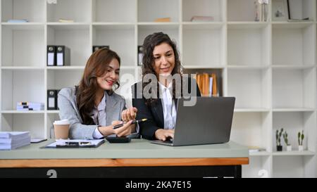 Two attractive millennial caucasian businesswomen business colleagues working together in the modern office, using and looking at laptop screen. Stock Photo