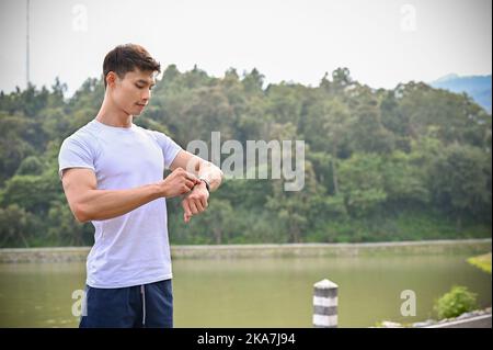 Handsome and fit young Asian athletic man in sportswear, checking his running miles and calorie burning on his smartwatch. Sport gadget concept Stock Photo