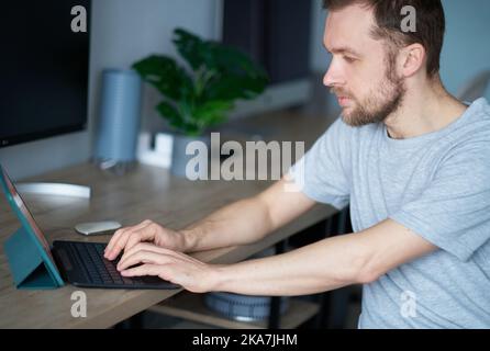 Cheerful bearded male software engineer sitting at working place in office hardly working using laptop. Young handsome man in grey t-shirt, positive emotion with computer monitor on background Stock Photo