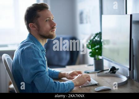 Cheerful bearded male freelance software engineer sitting at working place in home office hardly working using computer. Young handsome developer man in denim shirt. High quality image Stock Photo