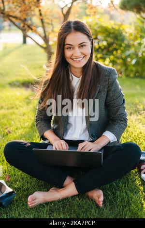 Young businesswoman student freelancer using laptop for distant remote work sitting on green grass lawn, looking at camera and smiling. Summer Stock Photo