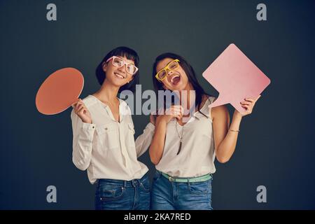 Guess whats on our mind. Studio shot of two cheerful young women holding up speech bubbles while standing against a dark background. Stock Photo