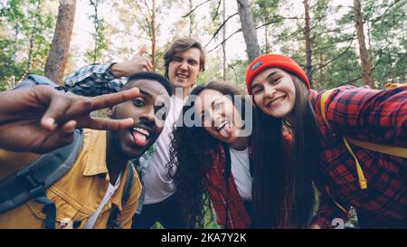 Multiracial group of friends travelers is taking selfie in wood looking at camera, posing with funny faces and trendy gestures. Millennials are wearing modern tourist outfit. Stock Photo