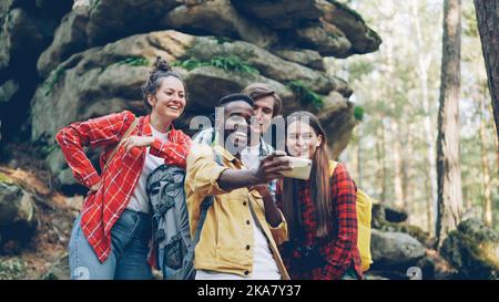 Funny young people happy friends are taking selfie in wood with mossy rocks in background, African American guy is holding smartphone, smiling men and women are posing. Stock Photo