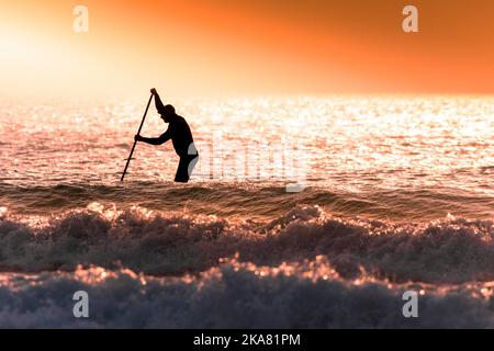 A paddle boarder silhouetted by a spectacular sunset at Fistral Beach at the end of the warmest day of the year in Newquay in Cornwall in the UK. Stock Photo
