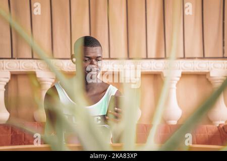 African American short haired man in true wireless earphones smiling while sitting on bench in cafe and browsing mobile phone Stock Photo