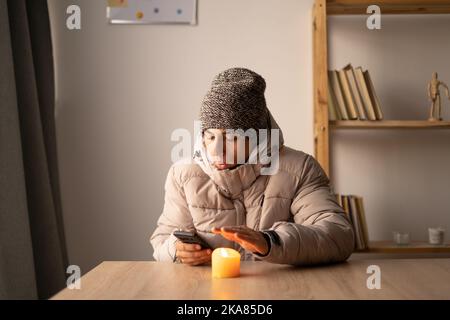 Young man warmly dressed is feeling cold, with warm jacket, sitting at the table at cold home. Have problem with health, central heating, energy Stock Photo