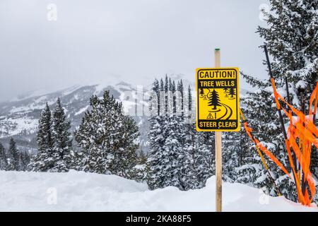 Sign warning of the danger of tree wells to skiers on top of snow-covered mountain. Stock Photo