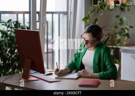 Focused Caucasian woman tutor or teacher making notes in notebook sits at table with computer Stock Photo
