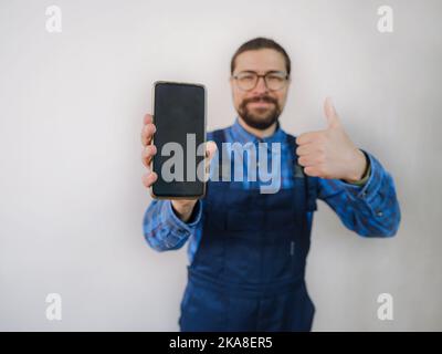 young man in blue work suit doing Repair apartment. Home renovation concept. Guy with smartphone. shows phone screen, mockup Stock Photo