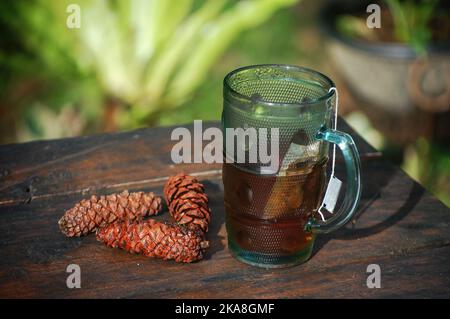 A shallow focus shot of a mug with hot tea in it placed on a wooden table with pine cones next to it Stock Photo