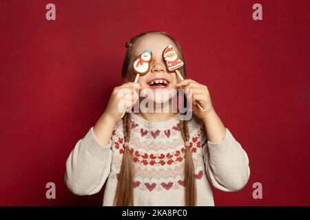 Funny joyful child girl having fun with Santa Claus and Snowman gingerbreads on red studio wall banner background Stock Photo