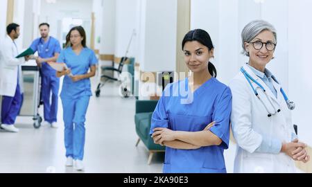Experienced chief physician posing with Asian nurse against background of medical staff walking along corridor of medical clinic. medical team Stock Photo
