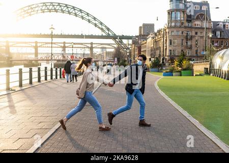 A young couple spending the day in Newcastle Upon Tyne. They are holding hands and walking near the River Tyne while the man looks back at his girlfri Stock Photo