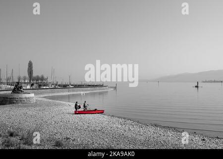A grayscale shot of the rocky beach of the sea with a red boat and passengers on the shore Stock Photo