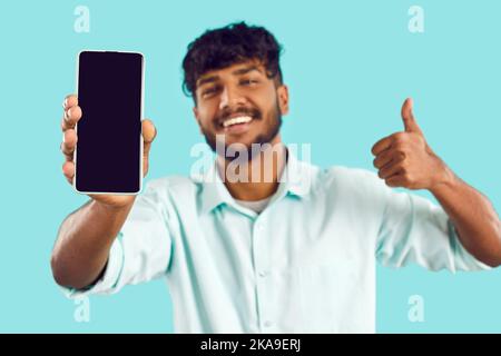 Smartphone with black blank screen in hand of happy man showing thumb up on light blue background. Stock Photo