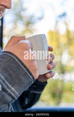 man's hand holding a cup of coffee next to a window with autumn background outside Stock Photo
