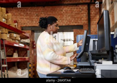 Side view portrait of adult black woman using computer while setting up printing machines in industrial shop Stock Photo