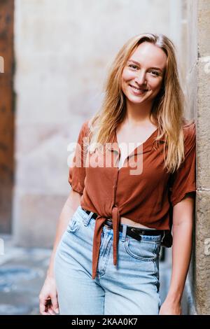 vertical portrait of a cheerful young caucasian tourist. She is leaning on a wall of old town and looking to camera Stock Photo