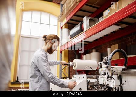 Side view portrait of young man putting in material in industrial printing machine at workshop, copy space Stock Photo