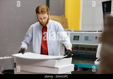 Portrait of young man working in print shop and holding stack of paper, copy space Stock Photo