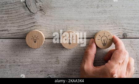 Male hand placing three wooden cut circles with light bulb icon drawn on them over textured white wooden background. Stock Photo