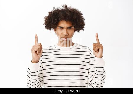 Offended hispanic guy, frowning upset, pointing fingers up, showing something bad, standing over white background Stock Photo