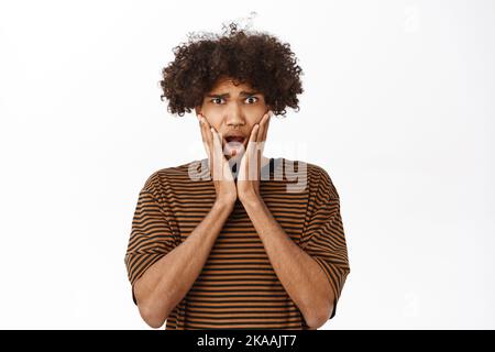 Portrait of hispanic guy looks with shocked face at camera, seeing smth terrible, reacting startled, standing over white background Stock Photo