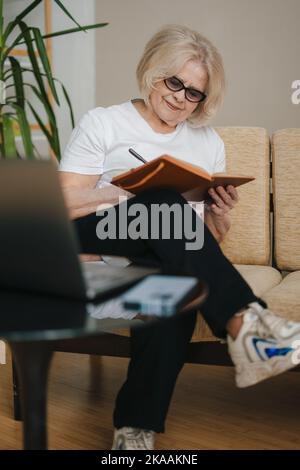 Happy senior woman watching online training webinar using laptop, remote working, writing notes. Senior woman using laptop while sitting on sofa at Stock Photo