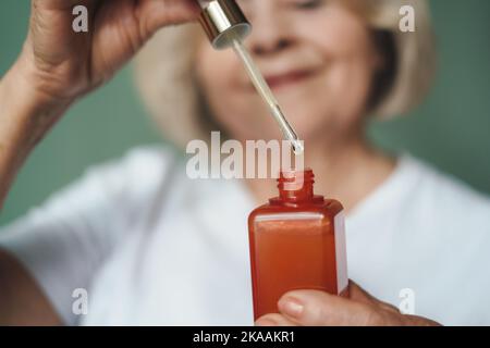 Portrait with focus on hands of senior woman holding bottle with oil collagen serum. Beauty procedure professional cosmetics usage at home. Protection Stock Photo