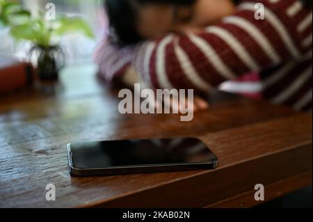 A smartphone is on a wooden table over blurred background of tired young Asian woman lay her head down or napping on the table. selective focus image Stock Photo