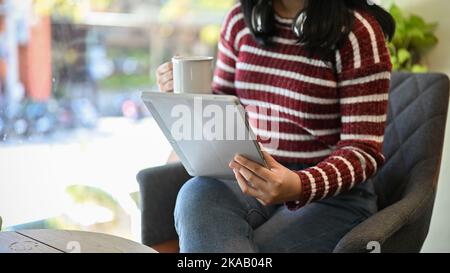 Beautiful young Asian woman in casual clothes sipping coffee and using her digital tablet while relaxing in the coffees shop. cropped image Stock Photo