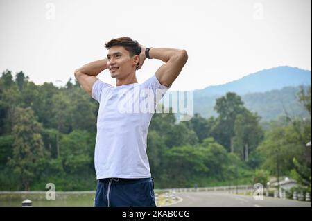 Handsome and sporty fit young Asian man in sportswear put his hands behind his head, taking a deep breath, resting after had a long run. Stock Photo