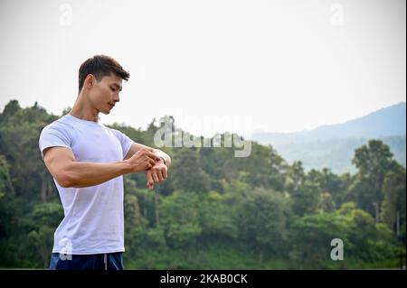 Sporty fit young Asian athletic man in sportswear checking his running miles on his smartwatch, running or jogging outdoors. Sport gadgets concept Stock Photo